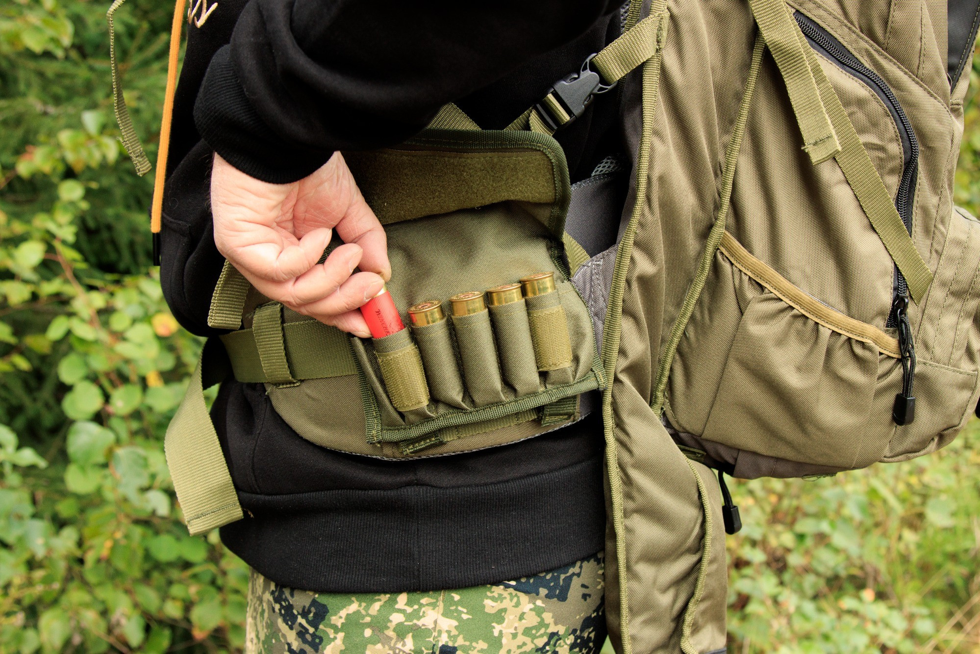 panostasku bear claw haulikolle-3 | Suomen Erämetsästys Person wearing a green tactical vest and camouflage pants places a red shotgun shell into a shell pouch on the vest.