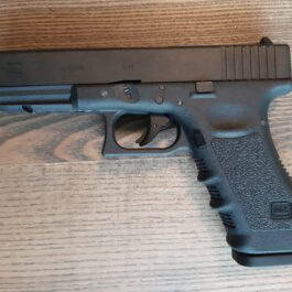 Black Glock-style handgun laid on a wood surface, showing the slide, trigger, and textured grip (side profile).