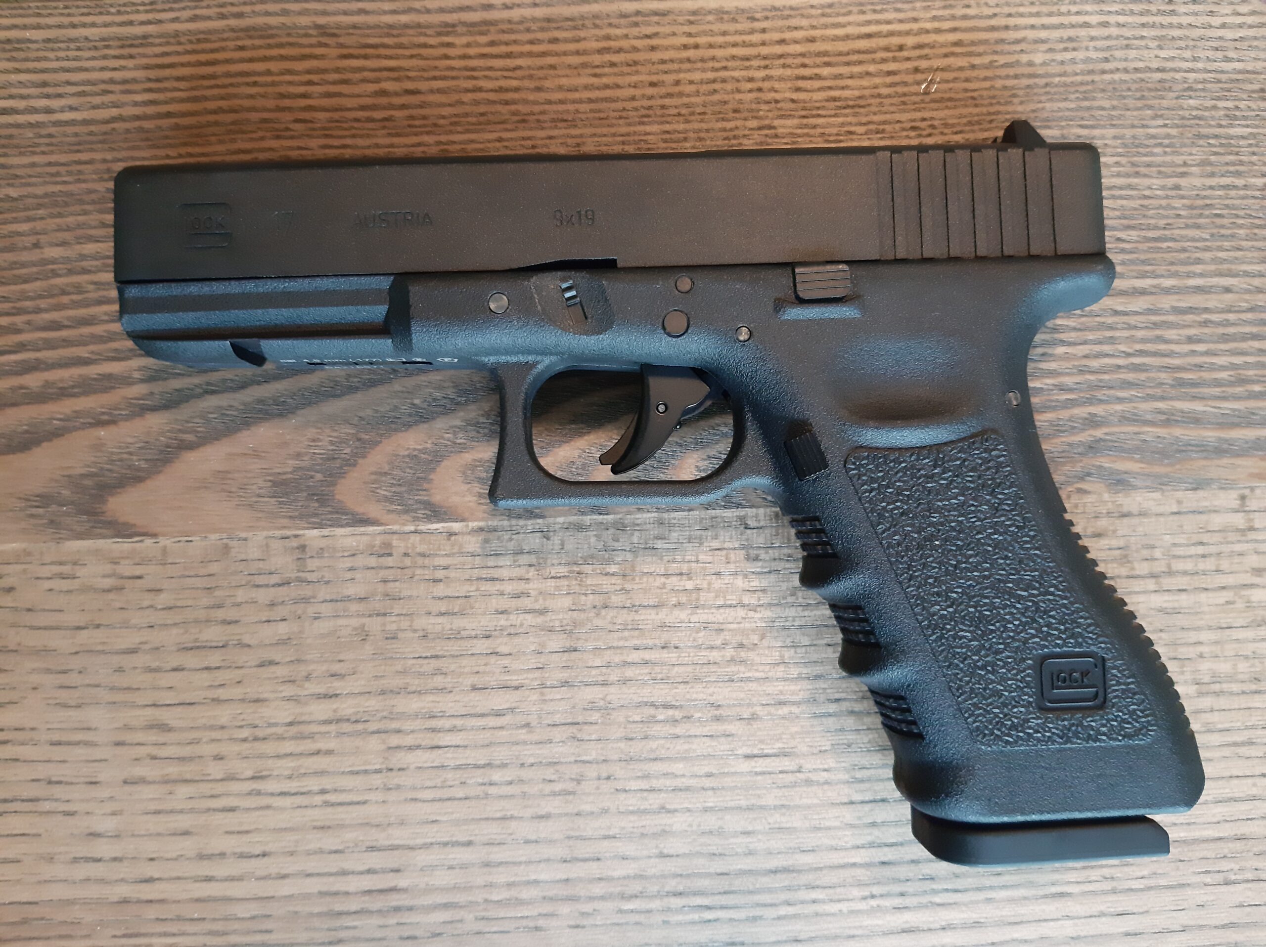 Ilmapistooli Umarex Glock17 Gen3 | Suomen Erämetsästys Black Glock-style handgun laid on a wood surface, showing the slide, trigger, and textured grip (side profile).